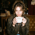 Woman holding a white mug with text in a cozy room.